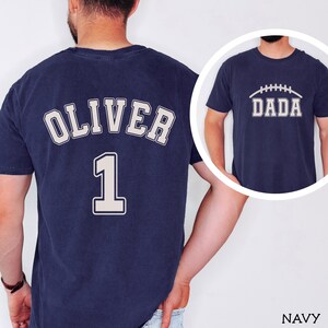 May include: A navy blue t-shirt featuring the name OLIVER and the number 1 in a light-colored collegiate font on the back. A circular inset shows the front of the same navy blue t-shirt with the word DADA and a football graphic printed on it.