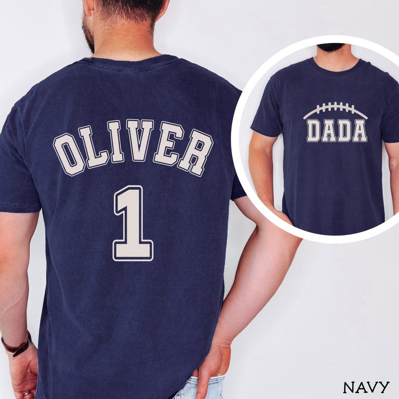 May include: A navy blue t-shirt featuring the name OLIVER and the number 1 in a light-colored collegiate font on the back. A circular inset shows the front of the same navy blue t-shirt with the word DADA and a football graphic printed on it.