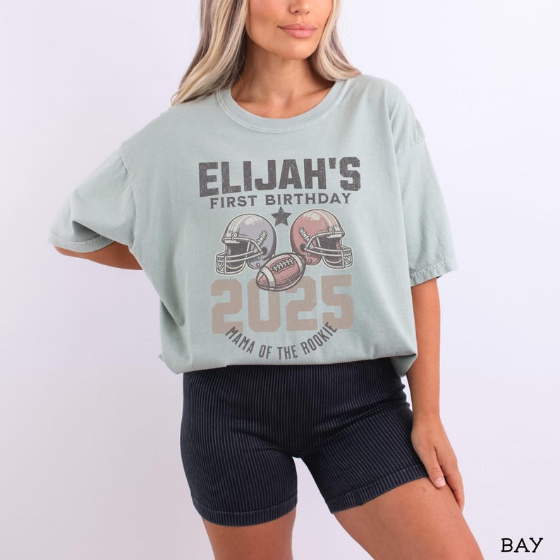 Puede incluir: Una camiseta de manga corta en color verde salvia claro con un gr&aacute;fico que incluye el texto ELIJAH'S FIRST BIRTHDAY 2025 MAMA OF THE ROOKIE, dos cascos de f&uacute;tbol americano y un bal&oacute;n. La camiseta se combina con pantalones cortos deportivos negros.