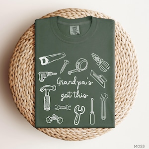 Peut inclure: Un t-shirt vert mousse avec des dessins au trait blancs d'outils, dont une scie, un marteau et une cl&eacute;. Le texte "Grandpa's got this" est &eacute;crit en police script. Le t-shirt est pr&eacute;sent&eacute; sur une surface circulaire tiss&eacute;e.