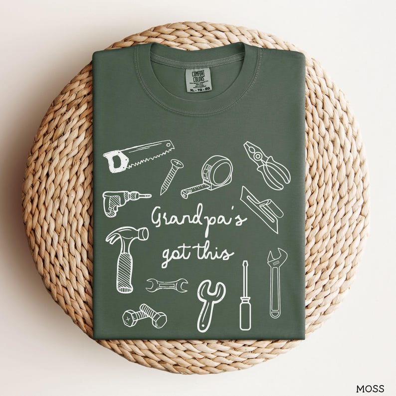 Peut inclure: Un t-shirt vert mousse avec des dessins au trait blancs d'outils, dont une scie, un marteau et une cl&eacute;. Le texte "Grandpa's got this" est &eacute;crit en police script. Le t-shirt est pr&eacute;sent&eacute; sur une surface circulaire tiss&eacute;e.