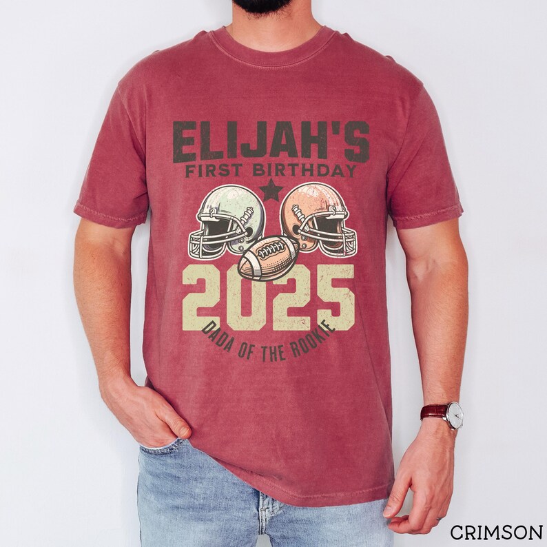 Puede incluir: Una camiseta de manga corta color carmes&iacute; con un gr&aacute;fico que incluye el texto ELIJAH'S FIRST BIRTHDAY 2025 DADA OF THE ROOKIE. El dise&ntilde;o presenta dos cascos de f&uacute;tbol americano y un bal&oacute;n de f&uacute;tbol en el centro, todo en estilo vintage.