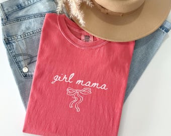 May include: A watermelon-colored short-sleeve t-shirt featuring the white text girl mama in a cursive script above a white line-art bow graphic. The shirt is displayed alongside light blue denim jeans, a tan wide-brim hat, and a gold necklace.