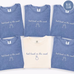 May include: Six blue jean-colored t-shirts with the phrase "last toast on the coast" and champagne flutes or a ring graphic. One shirt is cream-colored. The shirts are folded and displayed on a flat surface.