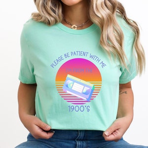 May include: A mint green t-shirt featuring a graphic of a VHS cassette tape centered over a circular sunset gradient of pink, orange, and yellow. The text reads, "PLEASE BE PATIENT WITH ME I'M FROM THE 1900'S" in a purple sans-serif font.
