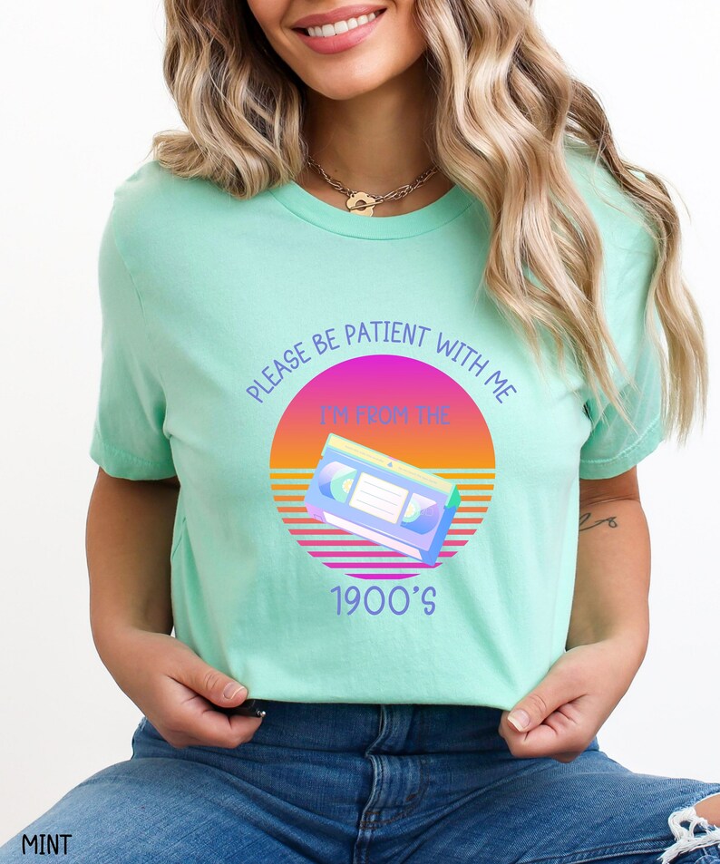 May include: A mint green t-shirt featuring a graphic of a VHS cassette tape centered over a circular sunset gradient of pink, orange, and yellow. The text reads, "PLEASE BE PATIENT WITH ME I'M FROM THE 1900'S" in a purple sans-serif font.
