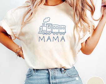 Puede incluir: Camiseta de color marfil con un gráfico de tren azul y la palabra "MAMA" en azul. El diseño incluye una locomotora y varios vagones. La camiseta está hecha de un material suave y es adecuada para el uso diario.