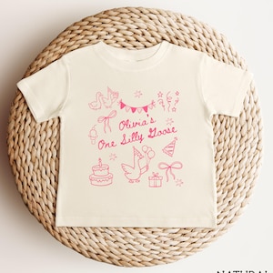 Op de afbeelding: Cr&egrave;mekleurig peuter-T-shirt met een roze afbeelding. Het ontwerp omvat een gans met ballonnen, een verjaardagstaart, feesthoeden, strikken en de tekst "Olivia's One Silly Goose". Het shirt wordt weergegeven op een gevlochten mand.