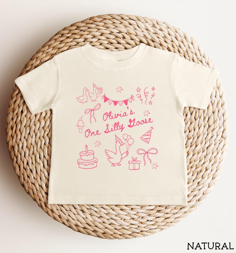 Op de afbeelding: Cr&egrave;mekleurig peuter-T-shirt met een roze afbeelding. Het ontwerp omvat een gans met ballonnen, een verjaardagstaart, feesthoeden, strikken en de tekst "Olivia's One Silly Goose". Het shirt wordt weergegeven op een gevlochten mand.