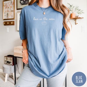 K&ouml;nnte beinhalten: Hellblaues T-Shirt mit dem Schriftzug "bun in the oven" und einer Grafik einer Zimtschnecke. Das Shirt hat eine lockere Passform. Der Text ist handschriftlich gestaltet. Das T-Shirt ist einfarbig.