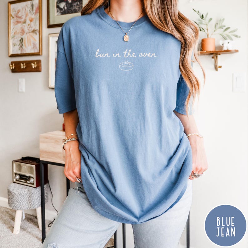 K&ouml;nnte beinhalten: Hellblaues T-Shirt mit dem Schriftzug "bun in the oven" und einer Grafik einer Zimtschnecke. Das Shirt hat eine lockere Passform. Der Text ist handschriftlich gestaltet. Das T-Shirt ist einfarbig.