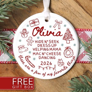May include: A round white ceramic Christmas ornament with a white ribbon hanger. The ornament features red line art illustrations of a gift, bow, snowman, tree, gingerbread man, car, wreath, candy cane, mug, and lights, with the text Olivia, 2026, and HIDE N' SEEK, DRESS UP, HELPING MAMA, MAC N' CHEESE, DANCING, these are a few of my favorite things.