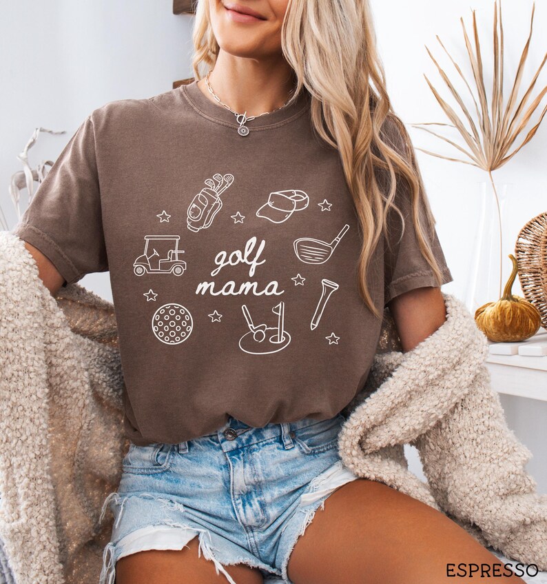 Puede incluir: Una camiseta marr&oacute;n con un dise&ntilde;o gr&aacute;fico blanco de iconos con tem&aacute;tica de golf y el texto "golf mama".