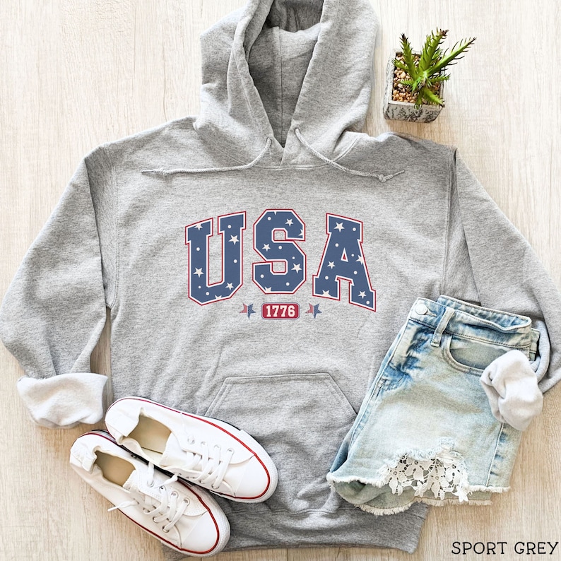 K&ouml;nnte beinhalten: Ein hellgrauer Kapuzenpullover mit dem Wort "USA" in blauen Buchstaben, gef&uuml;llt mit wei&szlig;en Sternen. Unter dem Wort steht die rote Zahl "1776". Der Hoodie wird mit Jeansshorts und wei&szlig;en Turnschuhen kombiniert.