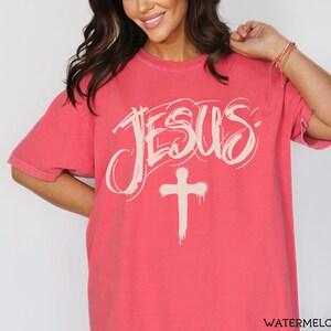 May include: A watermelon-colored t-shirt with the word "JESUS" in a stylized, off-white font above a matching cross. The shirt has short sleeves and a relaxed fit.