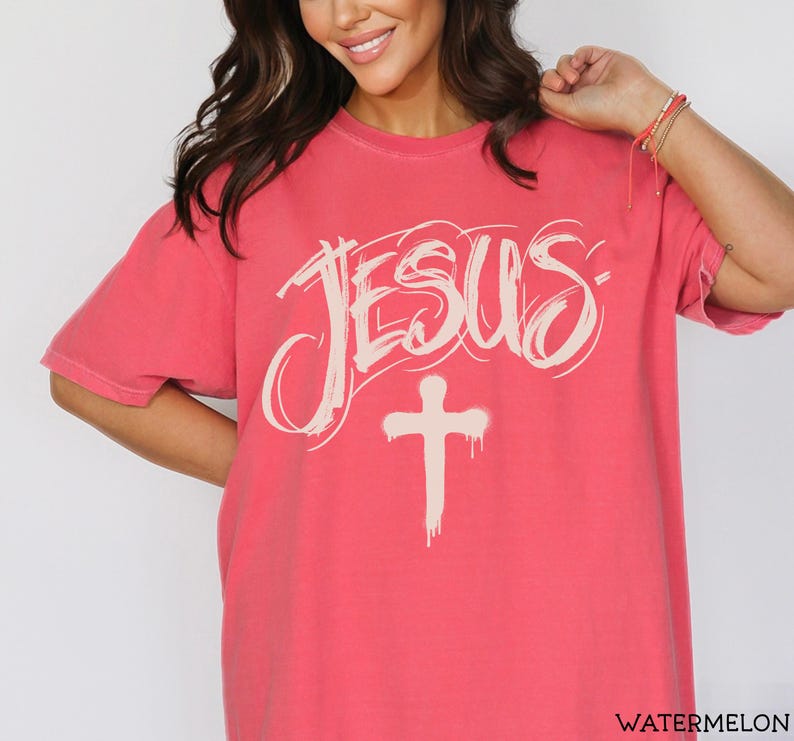 May include: A watermelon-colored t-shirt with the word "JESUS" in a stylized, off-white font above a matching cross. The shirt has short sleeves and a relaxed fit.