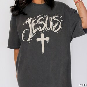 May include: Dark gray t-shirt featuring the word "JESUS" in a stylized, off-white script above a dripping cross. The shirt is a short-sleeved, crew neck style.