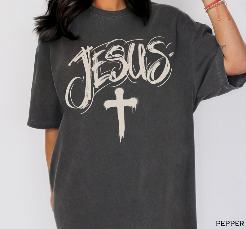 May include: Dark gray t-shirt featuring the word "JESUS" in a stylized, off-white script above a dripping cross. The shirt is a short-sleeved, crew neck style.