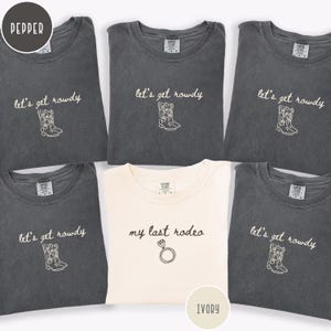 May include: Five pepper-colored t-shirts featuring the embroidered text let's get rowdy and a pair of cowboy boots, and one ivory t-shirt featuring the embroidered text my last rodeo and a diamond ring icon, all with crew necklines.