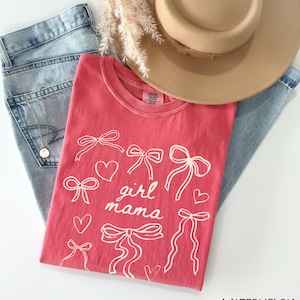 May include: A coral pink t-shirt with a white graphic design of bows and hearts. The text "girl mama" is centered on the shirt.