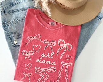 May include: A watermelon-colored t-shirt featuring white line art of various bows and hearts with the text girl mama printed in the center. The shirt is paired with light blue denim jeans, a tan wide-brimmed hat, and a delicate gold necklace.