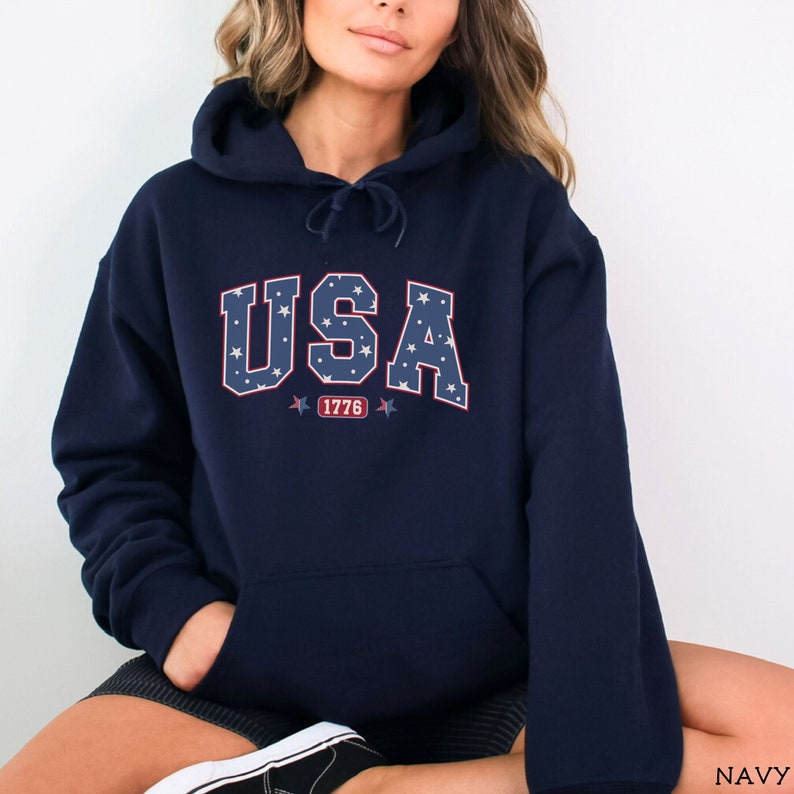 K&ouml;nnte beinhalten: Marineblauer Kapuzenpullover mit dem Wort "USA" in wei&szlig;en Buchstaben, gef&uuml;llt mit blauen Sternen. Die Zahl "1776" befindet sich unter dem Wort "USA" in einem rot-wei&szlig;en Design. Der Pullover hat eine Vordertasche.