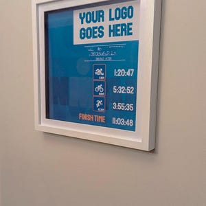 May include: A framed print with a white border displays a blue background with white text that reads "YOUR LOGO GOES HERE". Below are icons and times for swimming, biking, and running. The finish time is 11:03:48.