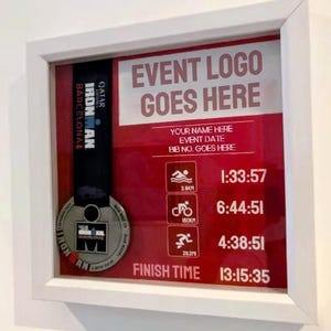 May include: A framed display featuring an Ironman Barcelona medal. The display includes a red background with event details, including swim, bike, and run times. The text "EVENT LOGO GOES HERE" is at the top, with a white frame.