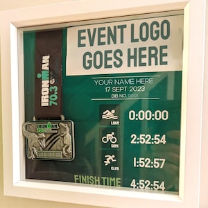 May include: A framed display featuring an Ironman 70.3 medal with a black and green ribbon. The display includes event details, a place for a name, and a finish time of 4:52:54. The text "EVENT LOGO GOES HERE" is visible.