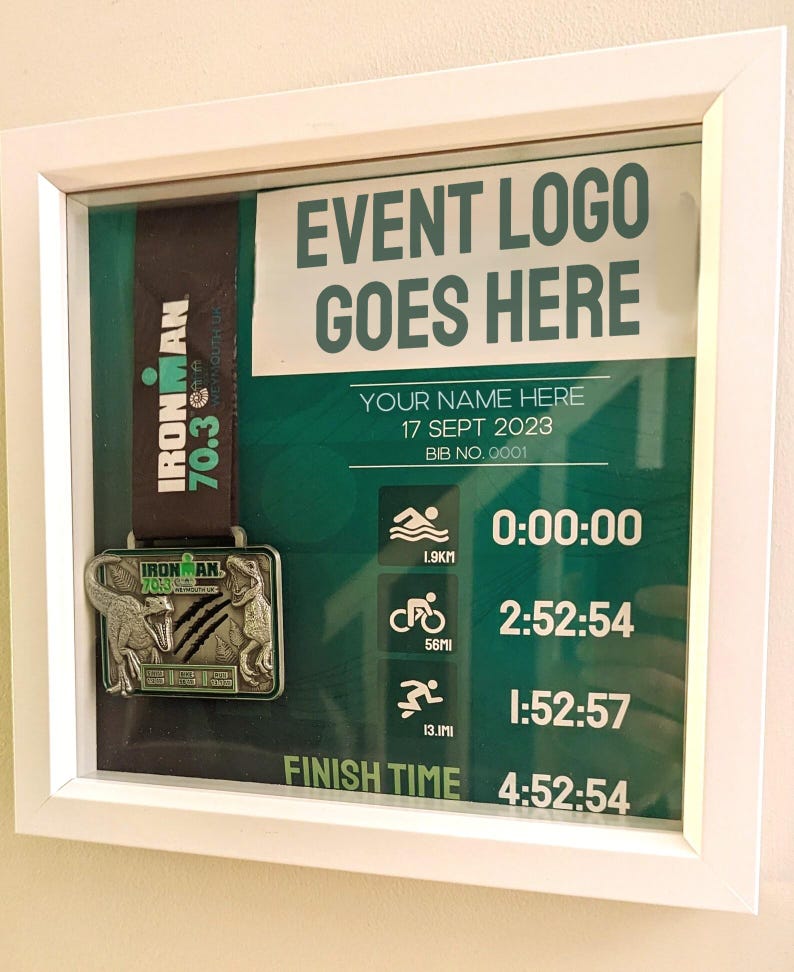 May include: A framed display featuring an Ironman 70.3 medal with a black and green ribbon. The display includes event details, a place for a name, and a finish time of 4:52:54. The text "EVENT LOGO GOES HERE" is visible.