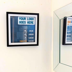May include: A framed print with a black border, featuring the text "YOUR LOGO GOES HERE" and race data. The print includes times for swimming, cycling, and running, with a total finish time of 13:22:59. The date is October 27, 2024.
