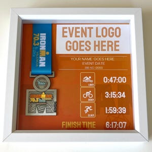 May include: A white framed display featuring an Ironman 70.3 Valencia finisher medal. The medal hangs from a blue ribbon. The display includes event details, a place for the event logo, and race times. The finish time is 6:17:07.