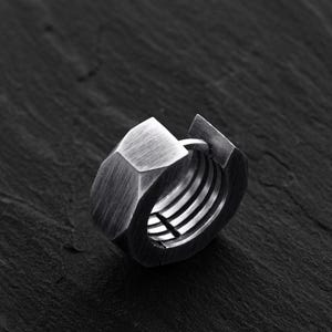Peut inclure: Une bague argentée de style industriel en forme d'écrou hexagonal. La bague présente un intérieur fileté détaillé et un design géométrique. La bague est posée sur un fond sombre et texturé.
