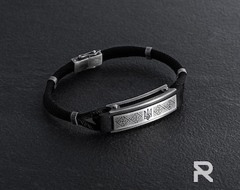 May include: A black braided bracelet with a silver rectangular plate featuring a Ukrainian trident symbol and intricate patterns.
