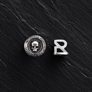 Könnte beinhalten: Ein silberner Anhänger mit einem Totenkopf und dem Text "Memento Mori Memento Vivere" sowie ein silberner Anhänger in Form des Buchstabens "R".