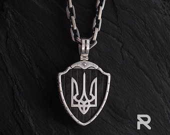 May include: A silver pendant necklace featuring a shield-shaped pendant with the Ukrainian Tryzub coat of arms. The pendant hangs from a silver chain. The background is a dark, textured surface.