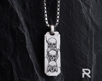 May include: A silver pendant necklace. The rectangular tag has a distressed finish and features three skulls, each with hands covering their eyes, ears, and mouth. The necklace has a square link chain.