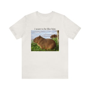 May include: A white t-shirt with a brown capybara sitting in grass with the text "i want to be like him" printed on the front.