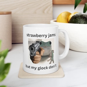 May include: White ceramic mug with a green frog holding a gun. The mug has the text "strawberry jams but my glock don't" printed on it.