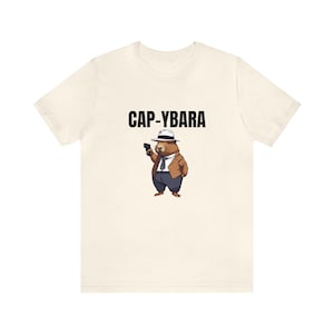 May include: A white t-shirt with a cartoon capybara wearing a fedora and holding a gun. The text "CAP-YBARA" is printed above the capybara.