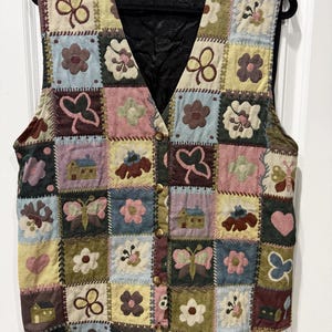Puede incluir: Un chaleco sin mangas con un diseño de patchwork de varios cuadrados con motivos bordados que incluyen flores, mariposas, corazones y casas pequeñas. El chaleco tiene un cuello en V, un forro interior negro y un cierre frontal con botones marrones.