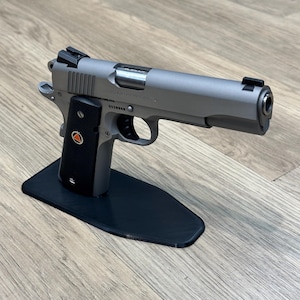 May include: A silver handgun with a black grip and a black stand. The gun has a silver slide and a black barrel. The stand is made of plastic and has a black finish.