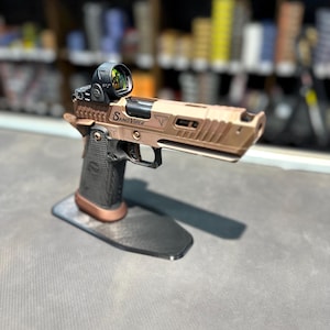 Puede incluir: Una pistola de color dorado con empuñadura negra y una mira de punto rojo. La pistola está etiquetada como "Sand Viper" y está en un soporte negro.