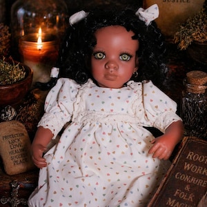 May include: A handcrafted doll with dark skin, black curly hair, and green eyes, wearing a white dress with a heart pattern. The doll is surrounded by candles, books, and other mystical items, suggesting a theme of spirituality and magic.