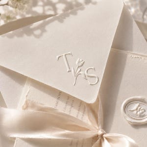 May include: Ivory-colored wedding invitation suite with embossed initials and a floral design. The suite includes an envelope, stationery, and a card sealed with a wax stamp. A cream-colored ribbon is tied around the stationery.