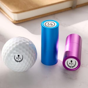 May include: A white golf ball with a black logo and two cylindrical stamps, one blue and one purple. The stamps have white logos on top. A brown notebook is in the background.