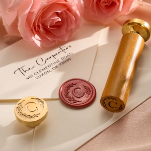 May include: A wooden wax seal stamp with a gold-coloured top and a wooden handle, next to a red wax seal and a gold-coloured wax seal. The envelope reads "The Carpenters" and their address. Pink roses are in the background.