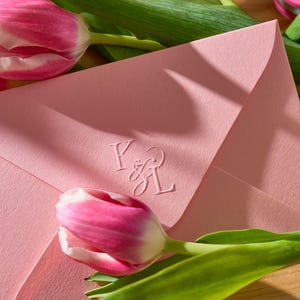 Könnte beinhalten: Ein hellrosa Umschlag mit geprägten Initialen, möglicherweise für eine Hochzeit oder ein besonderes Ereignis. Der Umschlag ist von rosa und weißen Tulpen mit grünen Stielen und Blättern umgeben. Die Szene wird von weichem, natürlichem Licht beleuchtet.