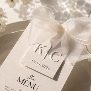 Peut inclure: Un menu de mariage crème avec un nœud en ruban blanc transparent. Le menu présente les initiales "K/C" et la date "12.19.2026". Le menu est sur une assiette blanche, avec des fleurs blanches en arrière-plan.