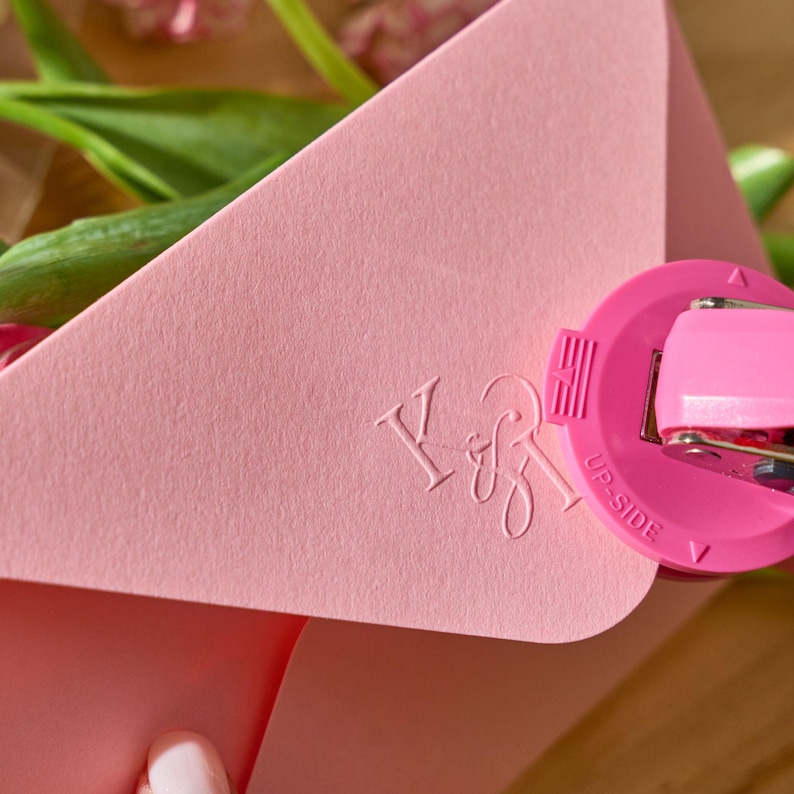 May include: A close-up of a pink envelope with embossed initials "K&B" and a pink stapler. The stapler is partially visible, with the words "UP-SIDE" printed on it. The background includes green leaves and pink flowers.